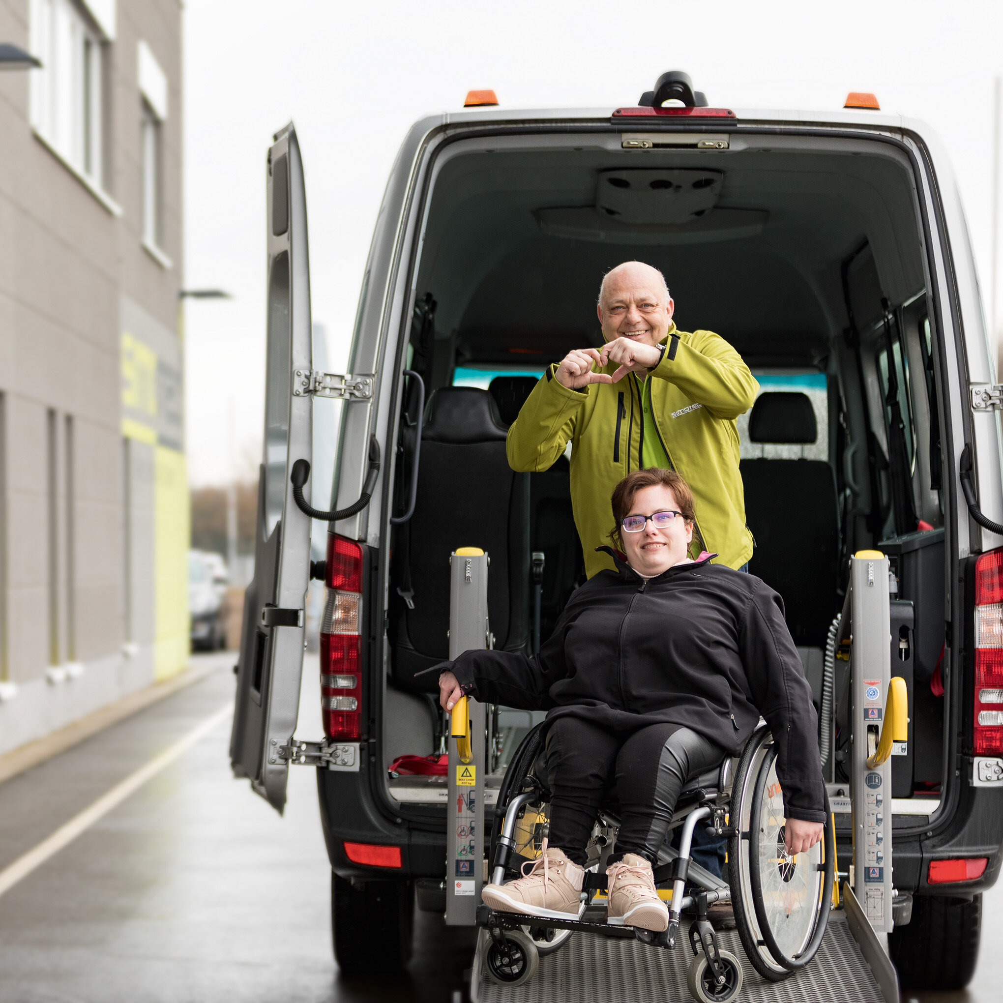 Eine Frau im Rollstuhl auf der Rampe eines Transporters und hinter ihr ein Mann der mit den Händen ein Herz formt